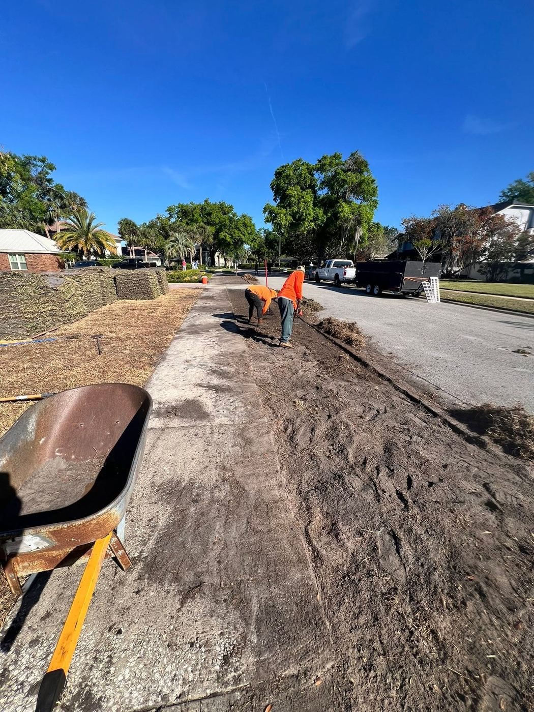 A cleared strip of bare graded soil running along a public sidewalk, with two workers raking it level and pallets of sod waiting on the left.